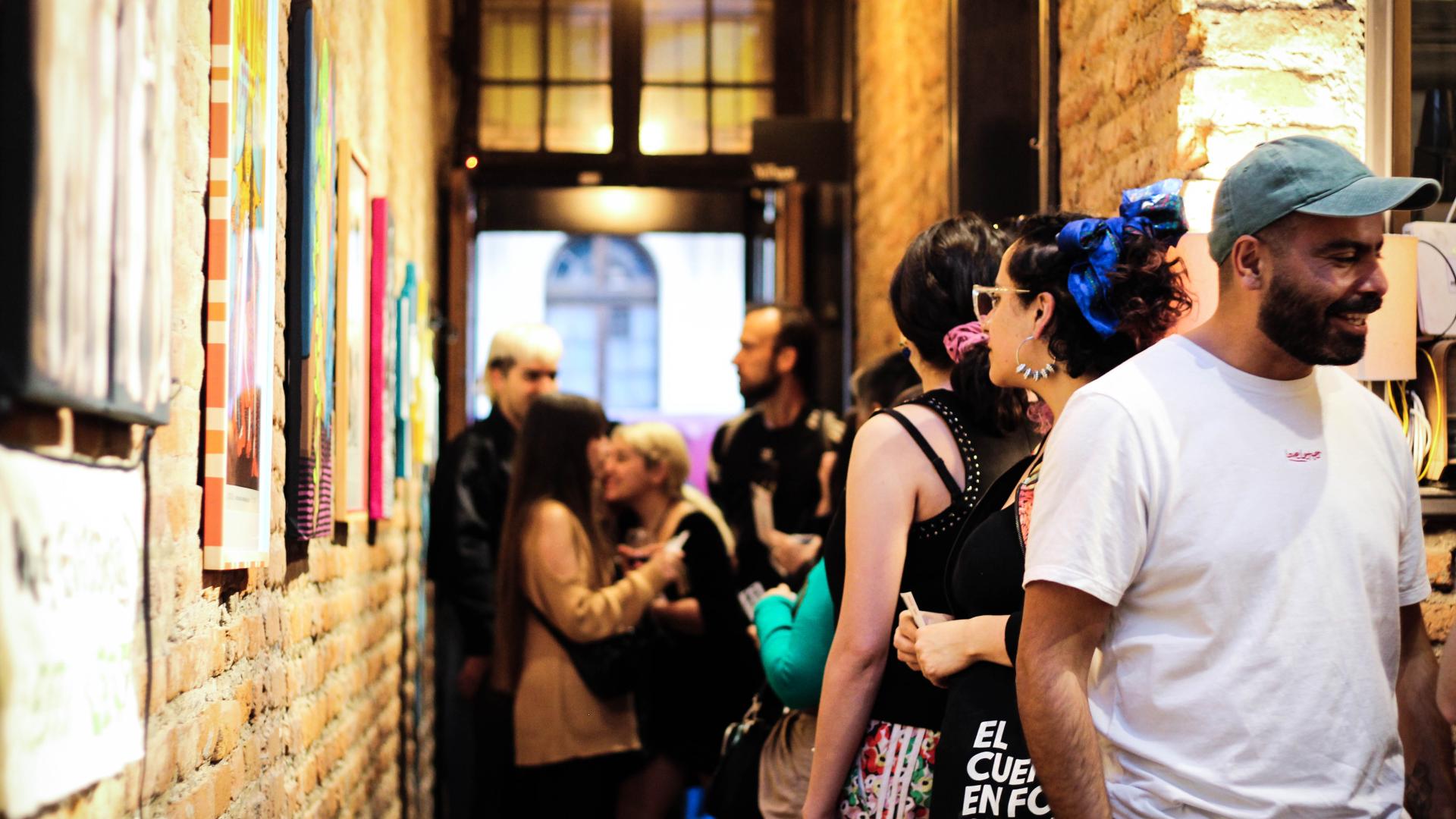 Vista desde el interior de Espacio 330, al costado izquierdo de la imagen el pasillo de ladrillo con las obras montadas, hay personas conversando y otras mirando la exposición