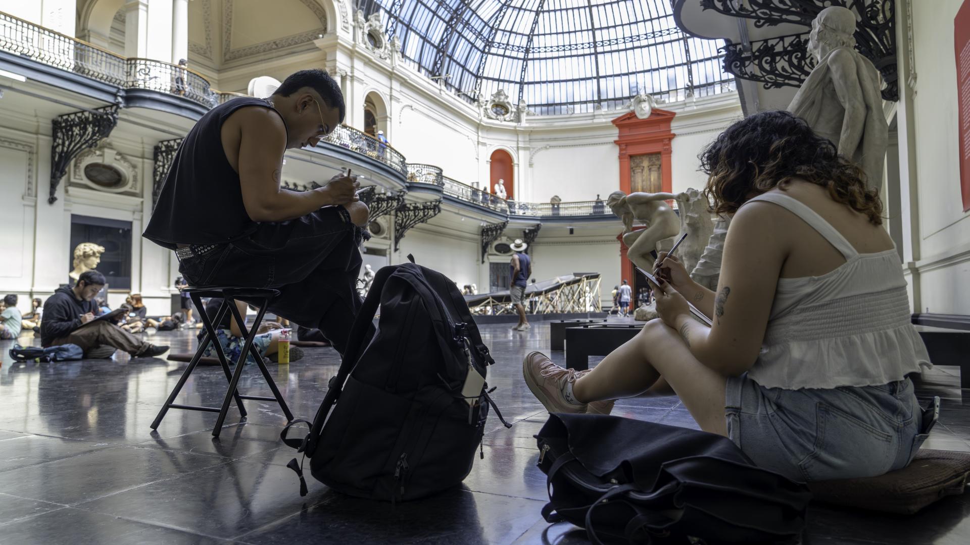 En el hall del Museo, dos personas en primer plano sentadas dibujando, se observa el espacio, parte de la cúpula vidriada y mas visitantes.