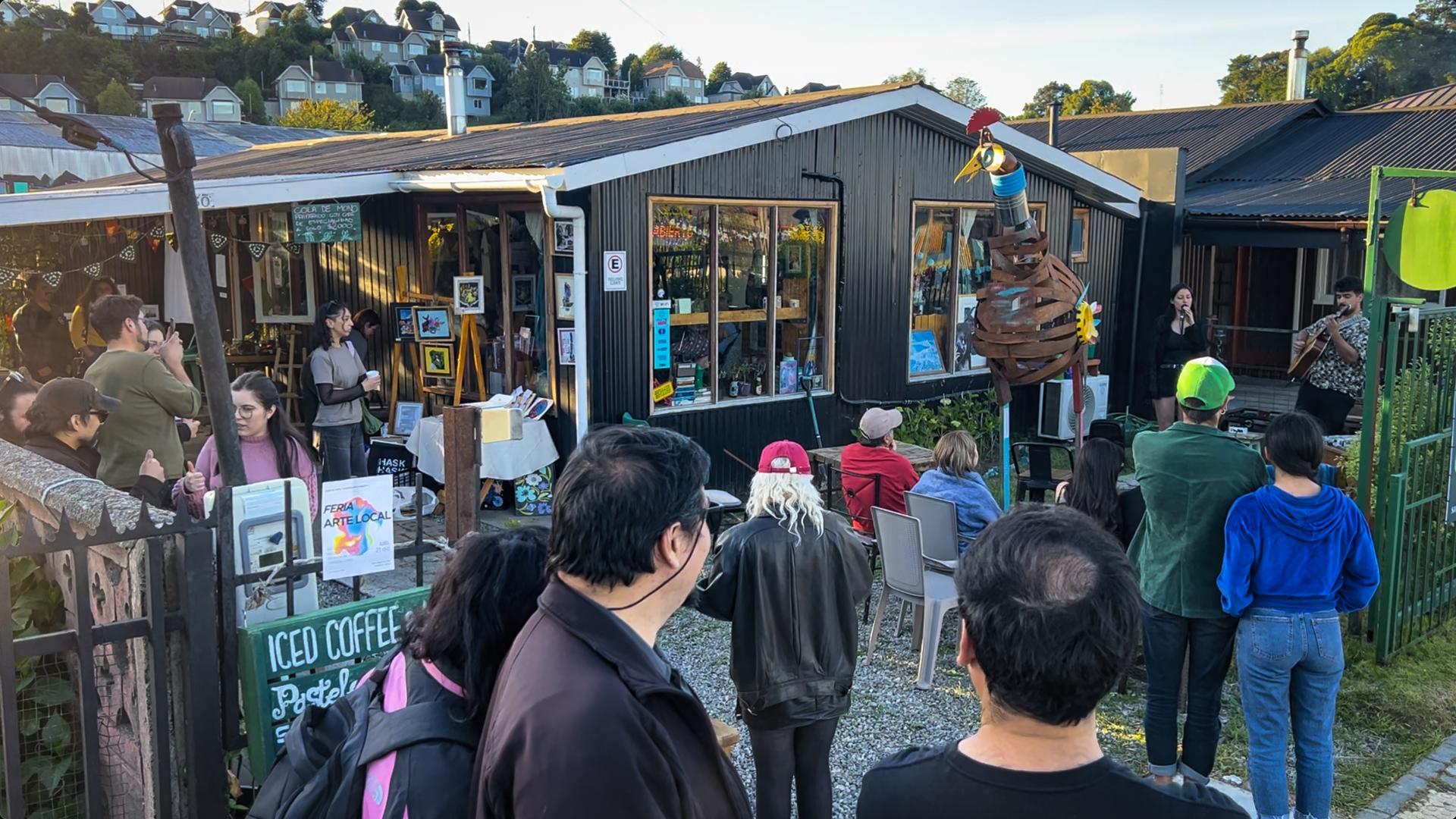Foto del exterior en el jardín de la casa Bisiesto, un grupo de gente se encuentra de pie viendo a un dúo de voz y guitarra tocar en vivo mientras cae la tarde. En el jardín también se pueden ver esculturas de materiales reciclados y distintas obras de arte pertenecientes a artistas locales.