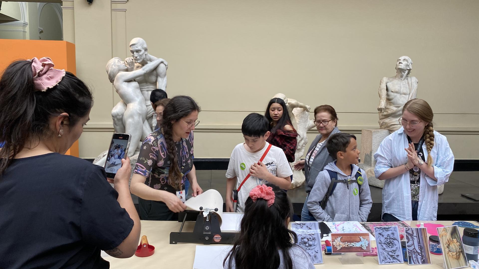Personas de adultas, una niña y dos niños en el hall del museo participando de la Estación de grabado, al fondo se ven parcialmente dos esculturas.