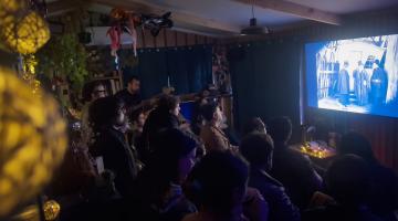 Unas 25 personas sentadas en una sala de microcine viendo el clásico de expresionismo alemán "El gabinete del doctor Caligari" (1920) con música en vivo, la sala está oscura y la luz de la pantalla es lo único que ilumina a la gente. 
