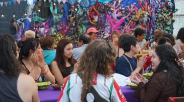 Personas conversando sentadas mientras comparten una cena. En el fondo una tela de muchos colores con materiales reciclados.